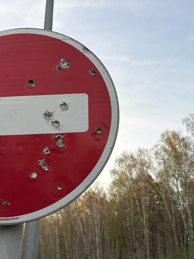 The image shows a damaged "No Entry" road sign, riddled with bullet holes. The sign is red with a white horizontal bar and is mounted on a metal pole. The background includes trees and a cloudy sky. The location of the image is specified as Таскино, Russia. The sign has been damaged by multiple shots, leaving holes and deformed metal.