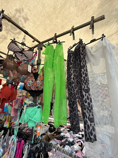 This is a medium shot, eye-level, outdoor photograph of a clothing stall, likely a market, under a beige awning. The stall displays a variety of swimwear and cover-ups hanging on metal racks. In the foreground, to the left, are several bikini tops and bottoms, some in solid red and green, others with patterned designs. Hanging centrally are a pair of lime green mesh pants and a matching bikini top, as well as a pair of black and white leopard print sheer pants. To the far right is a white lace-like sheer cover-up. The background shows more clothing items piled on the ground and hanging on racks, suggesting a busy retail environment. The overall scene is well-lit with natural light filtering through the awning.