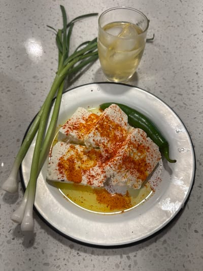 A top-down, close-up shot displays a prepared snack and drink on a speckled light grey surface, likely a kitchen counter. In the center, a white enameled plate with a black rim holds several irregular, rectangular blocks of white cheese, possibly feta or a similar soft cheese. These cheese portions are generously sprinkled with vibrant red paprika or chili powder and glistened with a drizzle of olive oil, which pools slightly at the bottom of the plate. A single, long green chili pepper rests beside the cheese on the plate.

To the left of the plate, a clear glass contains a light golden liquid with several ice cubes, indicating a cold beverage. The glass shows some condensation. Extending from the bottom left of the frame and angling towards the plate are several stalks of fresh green onions or scallions. The overall scene suggests a simple, rustic appetizer or mezze ready for consumption. There are no people visible in the image, nor is there any discernible text. The lighting appears to be artificial, typical of an indoor setting.
