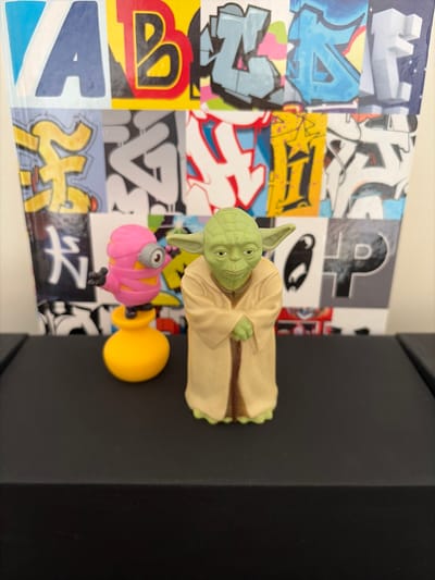 The image displays a close-up, eye-level shot of two toy figurines on a black surface. To the left, a pink-bandaged minion toy stands on a yellow, bell-shaped base. To the right, a Yoda figurine, in its typical beige robe and green skin, is positioned with its hands clasped in front. Both figures are facing forward. Behind them, a colorful collage of graffiti art on various panels serves as the backdrop. The graffiti features bold letters and abstract designs in a variety of colors, including blue, yellow, red, and white. The scene is well-lit, highlighting the details of the toys and the vibrant graffiti. The specific location in Malmö, Sweden is not identifiable from the image.