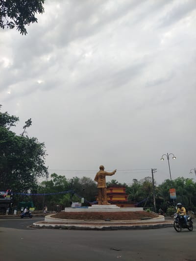 A daytime view shows a statue of a man in Chopdem, India. The statue is made of gold and stands on a platform, gesturing with its right arm. The area has been decorated with blue streamers. Trees surround the statue, and behind it is a structure with a red and yellow design. There is a road next to the statue, with a motorcycle carrying two people driving on it. Streetlights are visible in the background. The sky is overcast with a mix of gray and white clouds.