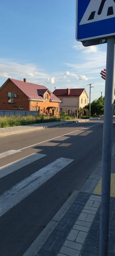 In Sokolivske, Ukraine, a pedestrian crossing is visible on a paved road under a bright blue sky with scattered clouds. Two houses with red roofs are situated behind a low fence to the left of the road. The house on the left is made of orange bricks, while the house on the right is lighter in color. A blue and white pedestrian crossing sign is mounted on a pole to the right of the frame, with a hand reaching towards it from the upper right corner. The sidewalk adjacent to the road is paved with interlocking bricks.