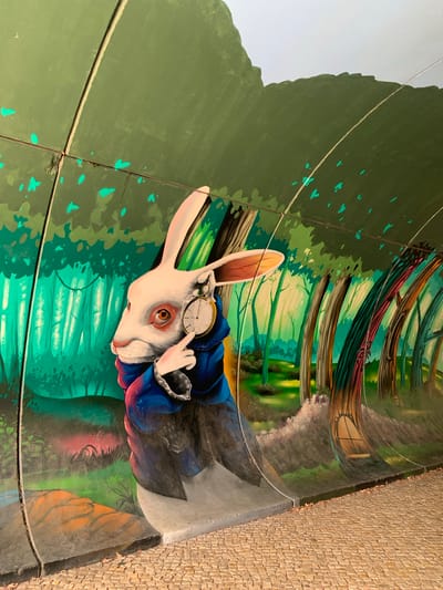 The image shows a mural of the White Rabbit from Alice in Wonderland, painted on the curved interior wall of a tunnel or underpass. The Rabbit is depicted in profile, wearing a blue coat and holding a pocket watch. It has large ears, reddish-brown eyes, and a pink nose. The background of the mural features a stylized forest with green trees and dappled light. The tunnel wall is made of segmented panels. The floor of the tunnel is paved with cobblestones. This mural is located in Odivelas, Portugal.