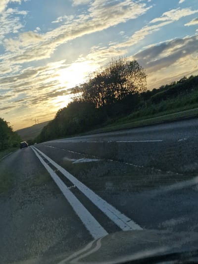 The image depicts a road with double white lines, adjacent to a grassy verge and a tree line silhouetted against a sunset sky with clouds. The road appears wet, possibly due to recent rain. A vehicle is visible on the road in the distance. The location is likely in a rural area, potentially near Witton Gilbert, United Kingdom, given the landscape. No people are directly visible in the frame.