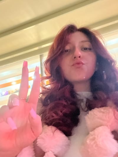 A female individual is depicted in a close-up, selfie-style photograph, taken indoors at an unspecified location within Piperkov Chiflik, Bulgaria. The subject has long, wavy, reddish-brown hair and is looking directly at the camera with her lips pursed. Her left hand is raised, with the index and middle fingers extended in a "peace sign" gesture. The fingernails on these fingers are long, rectangular, and light pink. A thin, gold-colored ring is present on the ring finger of the left hand. The individual is wearing a light-colored top, partially obscured by a fluffy, light pink or peach-colored outer garment with a distinct textured surface. The background is blurred, showing horizontal yellowish-beige structures at the top and indistinct orange and lighter strips below, consistent with an interior setting under artificial lighting.