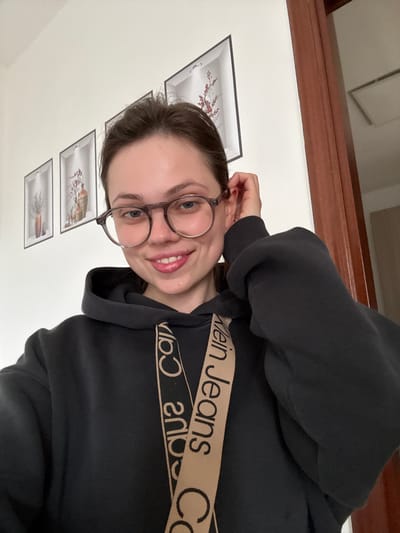 A female individual is depicted in an indoor setting, centered in a frontal, upward-angled selfie. The individual, appearing to be a young adult with brown hair, wears round clear-framed eyeglasses and a dark-colored hooded sweatshirt. A tan-colored strap, partially visible across her chest, displays reversed text, notably "Calvin Klein Jeans." Her right hand is positioned near her right ear, touching her hair or the frame of her glasses. She is smiling, revealing teeth.

The background features a plain white wall with three horizontally aligned framed pictures. Each frame contains an image of a vase with floral or botanical elements; from left to right, these include light purple sprigs, reddish branches, and white/red star-like designs. To the right of the subject, a dark reddish-brown wooden doorframe is visible, opening to an adjacent interior space with a white wall and a recessed light fixture or vent on the ceiling. There are no visual elements within the image to forensically confirm the specific location as Ercolano, Italy.