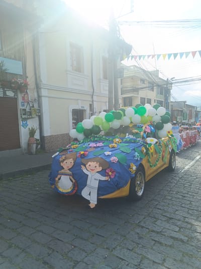 In Amaguaña, Ecuador, a street parade is depicted on a bright, sunny day with significant lens flare. The primary subject is a yellow compact car, extensively decorated. The car's front half is covered with a blue fabric adorned with various multi-colored cut-out flowers and green leaves. Two large cartoon-style cut-outs of children are attached to the front-left side: a girl in a white dress with red, yellow, and blue trim, and a boy in white clothing. The car's roof and rear are piled with white and green balloons, and its sides feature green garland-like decorations. The vehicle has shiny, possibly chrome, wheel rims and is positioned on a cobblestone street with grass growing between the stones.
Behind the decorated car, on the right side of the street, a procession of approximately ten young girls is walking in a line. Most are dressed in traditional attire, featuring white blouses and red skirts with white polka dots; one girl at the front wears a blue and white dress.
The left side of the scene features a narrow paved sidewalk alongside buildings. The closest building has a closed brown corrugated metal rolling door with partial signs for "REGALOS" (gifts) and "COPIAS" (copies) visible above. Adjacent to it stands a light-yellow building with white trim around its windows and corners. Utility boxes, electrical wiring, and signs for "Tuenti," "Movistar," and "banco banco aqui" are mounted on its exterior. Potted plants are present on the sidewalk.
The background on the right consists of additional multi-story buildings lining the street. Overhead, numerous black utility lines crisscross the sky, supported by poles, and colorful triangular pennant banners are strung across the street, indicating a festive atmosphere. The strong sunlight from the upper left creates significant lens flare, obscuring some details in the upper portion of the image.