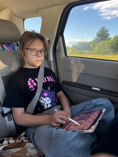 In a car, a young person with brown hair and glasses is lying down, looking at the camera. They are wearing a black t-shirt with a purple cartoon character and pink text. A seatbelt is fastened across their chest. To their right, another person in blue jeans is sitting and using a tablet with a stylus. The tablet screen shows a game with a pink checkered background. Outside the car window, trees and a cloudy sky are visible, suggesting a daytime scene. The interior of the car, with its beige seats and car door panel, indicates the setting is within a vehicle. The overall mood appears relaxed, possibly during a road trip. The scene is likely taking place in Бабаново, Russia.