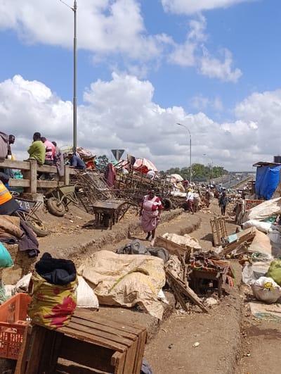 A cluttered scene depicts a makeshift market or settlement with debris, carts, and goods scattered along a dirt path. People are visible, some sitting on a bridge railing, while others walk through the area. The sky is partly cloudy, and a tall utility pole stands in the distance. The scene suggests poverty and informal economic activity.