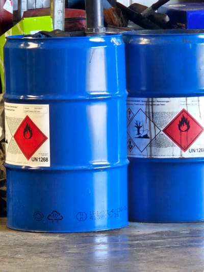 The image shows two blue industrial drums in what appears to be a storage area or workshop. The drum on the left is closer to the viewer and is mostly visible. It has a white label with red hazard symbols and text. Specifically, a red diamond-shaped label with a flame symbol and the text "UN 1268" is prominent. Below this, there are other hazard symbols, including one depicting a dead tree. The drum on the right is partially obscured by the left drum and shows similar labeling. Both drums appear to be made of plastic or metal and are cylindrical. The background is cluttered with various objects, including what looks like tools and other containers, suggesting a functional, possibly industrial or DIY, environment. The lighting is moderate, and the surface beneath the drums appears to be a concrete floor. The image is taken from a slightly low angle, looking up at the drums. The context provided about Bemmel, Netherlands, is external information not directly inferable from the visual content of the image itself.