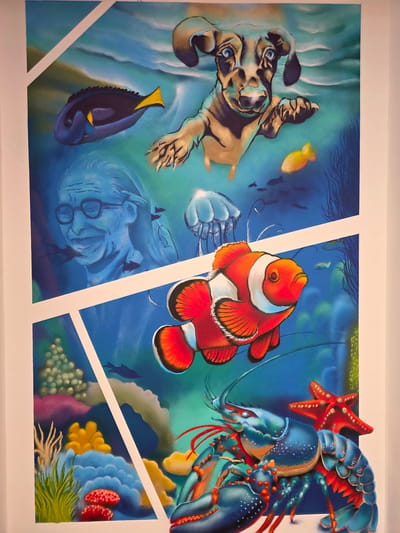 The image shows a large, colorful mural painted on a wall, depicting an underwater scene. The mural is divided into sections by diagonal white lines. In the upper section, a golden-brown dachshund dog appears to be swimming, with its paws outstretched. Below the dog, a blue tang fish swims to the left, and to the right of that, a small yellow fish. Further down, a portrait of a person wearing glasses and smiling, rendered in shades of blue, is visible. Small silhouetted fish are scattered throughout the upper portion of the mural.

The lower section of the mural features a vibrant clownfish swimming prominently, with red, white, and orange markings. Below and to the right of the clownfish, a large blue lobster with red accents is depicted. A red starfish is positioned behind the lobster. The background of this section shows various colorful coral formations and sea anemones in shades of green, yellow, blue, and red.

The mural is located in Halle (Saale), Germany, specifically at the "Marktschlösser" complex in Halle-Neustadt. The artwork is part of an art intervention project aimed at revitalizing the area.