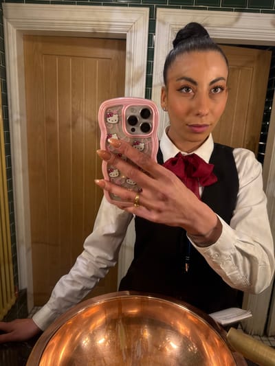 The image is a close-up selfie of a woman wearing a white button-down shirt with a black vest and a dark red bow tie. Her dark hair is pulled back in a bun. She is holding a pink smartphone case with Hello Kitty designs. A large copper bowl with reflective interior is visible on the right. In the background, there is a door and wall paneling with green tile accents. There is no definitive information to establish the location in Warrington, United Kingdom.