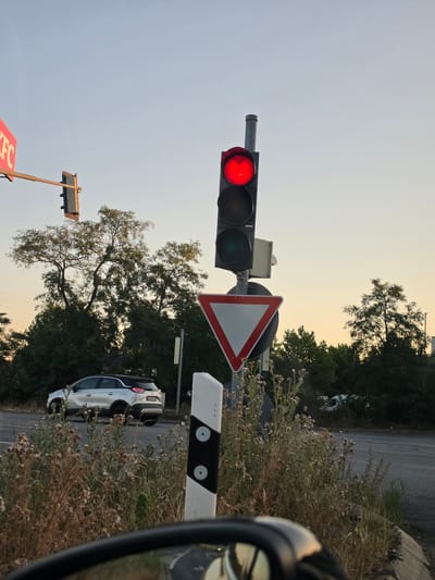 In Bischofsheim, Germany, a traffic light displays a red signal, indicating a stop. A white car is parked on the side of the road, partially obscured by overgrown vegetation. A yield sign is visible beneath the traffic light. The scene is set against a backdrop of trees and a clear sky, suggesting an outdoor urban environment.