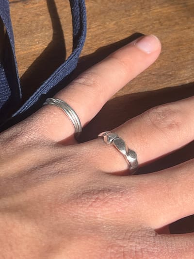 The image displays a close-up, high-angle view of a person's left hand resting on a wooden surface, located in the city of Hof Ashkelon Regional Council, Israel. The hand's skin tone is light, with visible skin texture and creases. Two silver-toned rings are worn on the hand: one on the ring finger, consisting of three thin, parallel bands that appear twisted or stacked; and another on the middle finger, featuring an undulating, patterned design with rounded, raised elements. Strong, natural light illuminates the scene, casting distinct shadows of the fingers and rings onto the wooden surface. A dark blue, woven fabric strap or cord is visible in the upper left portion of the frame, partially out of focus, lying on the wooden surface. The wooden surface itself exhibits a visible grain pattern.