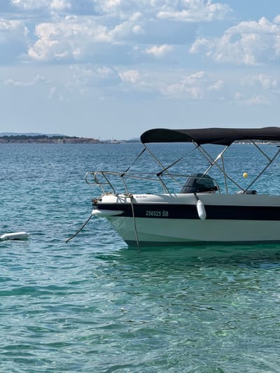 A white and dark blue motorboat is moored in the clear turquoise water of the Adriatic Sea, facing right. The boat has a black canopy supported by metal poles and a windshield. The registration number "256525 ŠB" and the text "Rent me:" are visible on the side of the boat. A white fender hangs from the side, and a white cylindrical buoy is floating in the water to the left of the boat. In the background, a coastal city with buildings and hills is visible across the water, likely within the administrative area of Grad Šibenik, Croatia. The sky above is blue with scattered white clouds.
