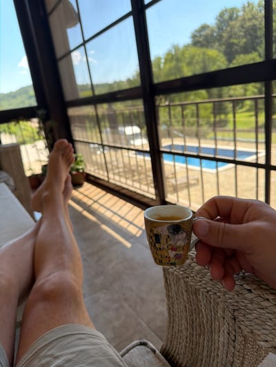 In Sokolovo, Bulgaria, a person's legs and feet are crossed, resting on a beige sofa. Sunlight streams through a large window, casting striped shadows on the floor. The person holds a small, ornate coffee cup, decorated with a depiction of Gustav Klimt's "The Kiss." Beyond the window, a clear blue swimming pool is visible, surrounded by lush green trees and hills. The scene suggests a relaxing moment on a sunny day, likely at a private residence or resort.