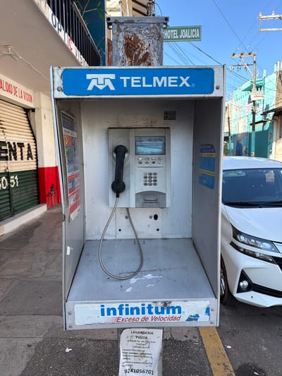 The image displays a Telmex payphone booth in a public area. The booth has a standard keypad, display screen, and attached receiver. The phone booth is showing signs of wear. Stickers and advertisements are present, including "infinitum" and a laundry service advertisement with a phone number. A street sign for "HOTEL JOALICIA" is also visible. A partial view of a white car is in the background. A shop is visible on the bottom left.