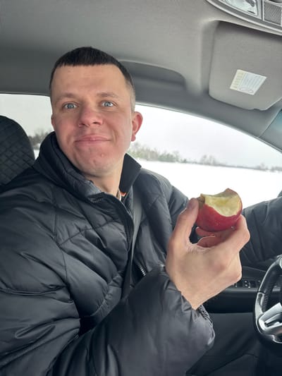 An image taken from inside a vehicle depicts a male occupant. The individual, appearing to be in the driver's seat, has short dark hair, light eyes, and is looking directly at the camera with a slight smile and raised eyebrows. He wears a black quilted puffer jacket, zipped up, with a black seatbelt visible across his chest. His right hand holds a red apple with a significant bite taken from it, exposing the white flesh. The vehicle's interior features a light grey headliner and sun visor, with a sticker containing Korean text visible on the visor. A dark car seat with a patterned headrest is partially visible behind him, and a dark steering wheel is to his lower right. Through the car window, a snowy, overcast winter landscape is visible, featuring snow-covered ground and distant trees. No specific identifying markers are present in the image to confirm the location as the city of Crokhovo, Russia.