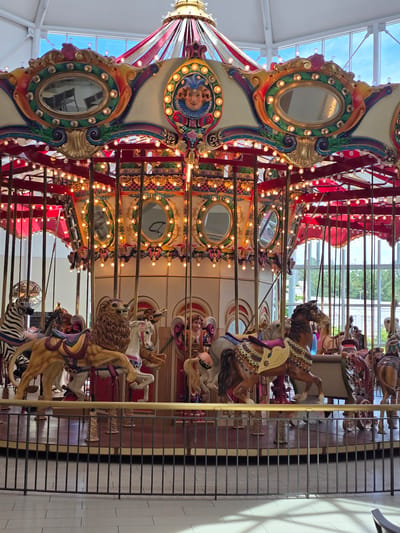 A carousel is depicted in a mall in Alpharetta, Georgia. The carousel is ornately decorated with gold trim, mirrors, and colorful paintings. It features several carousel animals, including a lion, a zebra, and multiple horses, each adorned with saddles and other embellishments. The carousel is illuminated by numerous small lights. In the background, a large window allows a view of the sky and trees outside, suggesting an indoor setting within a shopping center. The carousel is enclosed by a black metal fence.