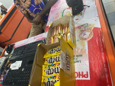This image provides a close-up view of a checkout counter in what appears to be a retail store, likely located in Goderich, Sierra Leone. A person is partially visible behind the counter, wearing a blue and brown patterned shirt and tan or khaki pants. Their hands are clasped together, suggesting they are a cashier or a customer interacting with the payment process.
The counter is equipped with standard point-of-sale hardware, including a black computer keyboard, a black mouse, a small black laptop with a stack of white receipts on it, and a black barcode scanner. To the left, a white receipt printer or similar device is visible.
Two cardboard display boxes filled with candy are positioned on the counter. The upper box contains numerous gold-wrapped "Twix" candy bars. Below it, the second box holds multiple yellow-packaged "M&M's" candies, with the brand name clearly visible. A small label on one M&M's package shows the number "10."
The counter surface is partly covered by a red and white mat or display. This surface features a cartoon lion face and various printed texts. Visible text includes "DNAO SAS - Capital 26 642 148 € - RCS Lyon 517 441 820 - Limonest - B," which pertains to a French company. Other upside-down words like "PHO," "Milk," "Lact," and Arabic script "واكه" (likely 'fruits') are also discernible, possibly from product advertisements on the mat. The overall scene depicts a functional retail transaction area.