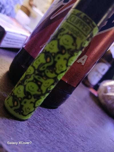 The image shows a green and black lighter, with a design of small smiling faces, lying on top of a brown glass bottle, partially obscured. The bottle label is red with white text. The background is out of focus but appears to be a dark wooden surface. The lighter and bottle are the only objects in focus. The scene is located at coordinates 50.439, 7.819 in the city of Montabaur, Germany. The objects are positioned on a surface that appears to be a table top. There is no indication of people in the image.