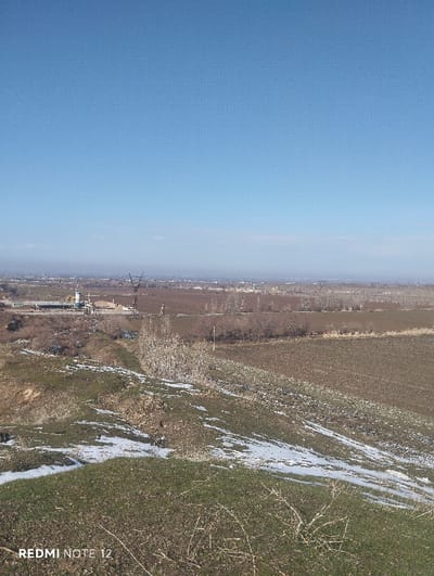 A landscape with a clear blue sky dominates the upper half. Below the horizon, a blurred cityscape fades into the distance. The mid-ground shows fields of brown earth and scattered trees, with a glimpse of industrial structures. The foreground presents a grassy hillside dotted with patches of snow. Dry vegetation and bare branches are visible, adding texture to the uneven terrain. The scene conveys a sense of winter's lingering presence on the land.