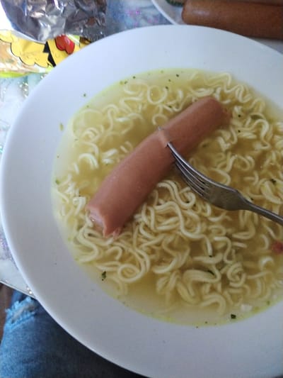 The image shows a close-up of a white bowl filled with instant ramen noodles and broth. A single hot dog, pierced by a silver fork, rests diagonally across the noodles. The broth is a pale yellow, with flecks of green herbs visible. To the upper left of the bowl, part of a foil wrapper with yellow and black markings is visible, along with some decorative sparkly material. In the background, another hot dog is partially visible. The setting appears to be a casual meal, possibly at home. The lighting suggests it could be daytime. The overall impression is of a simple, prepared meal. There is no visible text in the image.