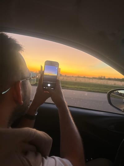 Inside a car, a person holds a smartphone to capture a sunset. The phone screen mirrors the outside view of the golden sky and adjacent landscape. The person's arm, wrist, and part of their face are visible, along with glasses. The car's interior is shadowed, contrasting with the bright light from the window. A side mirror can be seen reflecting the outside. The road and distant scenery are in the background.