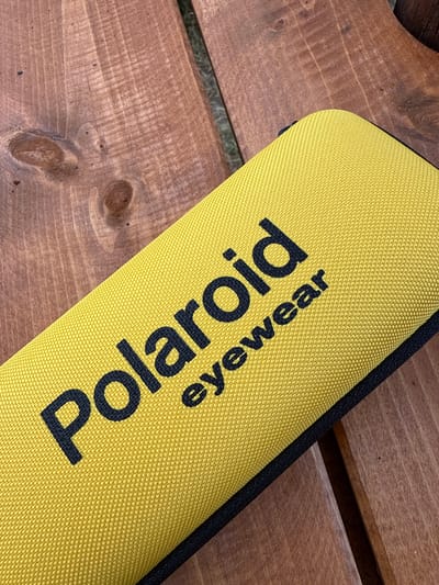 A bright yellow zippered case with "Polaroid eyewear" printed in black is placed diagonally on a weathered wooden surface. The wood has visible grain patterns and knots. The overall scene suggests an outdoor setting, possibly a park bench or a picnic table in Zgorigrad, Bulgaria. There are no people or actions depicted in the image.