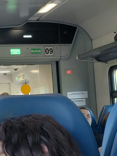 This media file depicts the interior of a train car in Russia. The primary subjects are two individuals, a person with dark hair on the right side of the frame and another person partially visible in the upper right, leaning back against a blue seat. The scene is a narrow train aisle with rows of seats.
Notable objects include a luggage rack above the seats, a sign with the number "09" and the word "ВЫХОД" (EXIT) in green lettering, a red illuminated sign with "WC" indicating a restroom, and a small, bright yellow circular object, possibly a button or a marker, on the right side of the frame. There are also several white rectangular signs with text in Russian, likely informational or cautionary. The lighting appears to be artificial, suggesting it is either daytime with limited natural light or nighttime.
The activity appears to be travel, with passengers occupying the train. The posture of the visible person suggests they are resting or sleeping. The overall atmosphere is utilitarian, typical of train interiors.
The visible text includes "09", "ВЫХОД", and "WC". Other signs have Russian text which is partially obscured or too small to read clearly. The location cue of "Kabinetnoye, Russia" provides context for the setting.