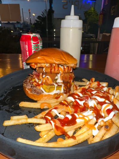 On a black plate sits a burger with fries, both liberally drizzled with multiple sauces, likely mayonnaise, ketchup, and possibly a cheese sauce. The burger is loaded with ingredients, including a beef patty, bacon, and onion rings. Beside the plate are squeeze bottles, presumably containing the sauces used on the burger and fries. A Coca-Cola can is also visible. The scene appears to be in a restaurant in Mérida, Mexico, judging by the reflected cityscape visible in the window. The time of day seems to be evening or night.