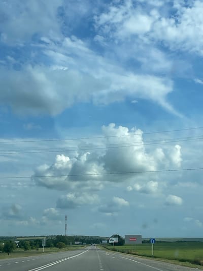 This media file depicts a scenic view from a vehicle traveling along a highway in Donskoye, Russia. The perspective is from inside the car, looking out at the landscape.
The primary subjects are the expansive sky filled with dynamic clouds, and the road stretching into the distance. The sky is a mix of bright blue and dramatic grey, indicating a day with varied weather, possibly a mix of sun and impending rain. Towering cumulonimbus clouds are visible, creating an impressive natural spectacle. In the foreground, power lines stretch across the frame, a common sight along highways.
The setting is a rural highway with a wide, paved road. To the right of the road, there is a green, cultivated field that extends to a line of trees. Further in the distance, more fields and trees are visible, suggesting a predominantly agricultural or natural landscape. There are also signs visible along the roadside. One sign is a large billboard with red text on a grey background. Above it, a blue sign with a white arrow indicates a turn. Another smaller sign is visible further down the road. A tall radio tower is also present in the distance.
The activity taking place is travel by road. The image captures a moment during a journey, showcasing the environment along the route. The time of day appears to be daytime, given the illumination and sky conditions. The overall mood conveyed is one of open space and the vastness of nature juxtaposed with human infrastructure.