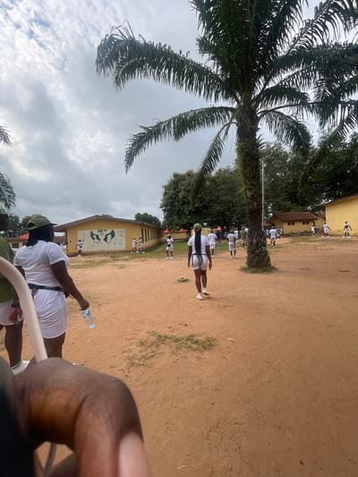 The image depicts an outdoor scene, likely in a rural or semi-urban area, characterized by reddish-brown dirt ground and scattered vegetation. Several individuals, predominantly young women, are present, all wearing white uniforms and green caps. They appear to be participating in some form of organized activity or gathering.

In the foreground, a woman in a white dress and green cap is seen from behind, holding a clear plastic water bottle. To her left, another person's arm and shoulder are visible, also wearing a green cap.

In the midground, a yellow building with "UNITY" and a graphic of wings painted on its facade is visible. Several individuals in similar white uniforms and green caps are scattered around this building and in the surrounding open area. Some are walking, while others appear to be standing or observing.

The background features more buildings, lush green trees, and a cloudy sky with palm trees dominating the upper right portion of the frame. The overall atmosphere suggests a sunny day, possibly in a tropical climate. The presence of the yellow building with the "UNITY" inscription and the uniformed individuals suggests a school, training facility, or a community event.

The location, Emure-Ekiti, Nigeria, is known for its educational institutions and agricultural activities. Given the attire and the setting, it is plausible that this image captures a scene from the National Youth Service Corps (NYSC) orientation camp in Emure-Ekiti, where young Nigerian graduates undergo a mandatory year of service. The presence of multiple young adults in uniform, a common sight in such camps, along with the building that could serve as a dormitory or administrative block, supports this interpretation.