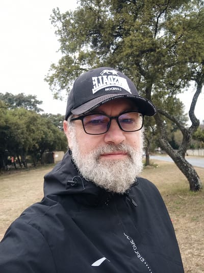 A medium shot portrait of a man with a full grey beard and glasses is captured outdoors. He is wearing a black hooded jacket and a black baseball cap. The cap has white text on it that appears to say "LONSDELE LONDON". The background consists of trees and dry grass, suggesting a natural or park-like setting. A paved road and fencing are visible in the distance. The lighting indicates an overcast or cloudy day. This image was taken in Randburg, South Africa.