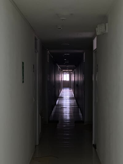 A long, narrow, dimly lit hotel hallway extends into the distance, terminating in a brightly lit window at the far end. The walls of the hallway are a pale, textured white, and the floor is tiled. Doors are visible on both sides of the hallway. On the ceiling, there are white circular smoke detectors and a white rectangular emergency light fixture. A green sign with a white pictogram is affixed to the left wall. The hallway appears to be empty of people. The overall impression is one of desolation and quiet. The image is undated and the location is unknown.