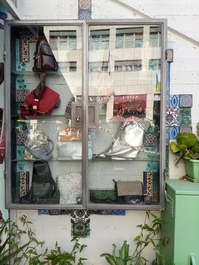 The image depicts a display cabinet filled with various handbags and decorative tiles, situated on a white, textured wall. The cabinet has two glass doors, revealing its contents. On the top shelf, a brown leather crossbody bag is hung, and a red handbag with white floral embellishments rests below it. The middle shelf displays a silver, shimmering bag and a pale blue, textured bag. The bottom shelf features a black backpack, a floral-patterned clutch, and a woven clutch bag. The cabinet is surrounded by decorative tiles with intricate patterns in blue, green, and red hues. In the lower part of the image, green foliage and plants are visible, suggesting an outdoor setting. A light green metal cabinet or storage unit is positioned to the right of the display cabinet. Reflections of buildings with windows are visible in the glass doors of the display cabinet. The scene appears to be a shop window display in Be'er Sheva, Israel, likely on Keren Hayesod Street, given the architectural style and the presence of similar displays in that area.