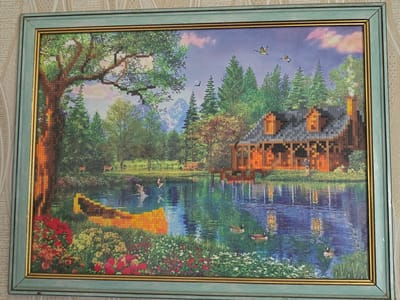 This diamond painting depicts a serene lakeside scene with a log cabin. In the foreground, lush green grass and vibrant red and yellow flowers border the water's edge. The lake is calm, reflecting the sky and the surrounding trees. Several ducks are visible on the water, with one in the process of taking flight from the water's surface.
In the middle ground, a rustic log cabin with a dark roof and glowing windows sits nestled among tall green trees. The cabin's reflection is clearly visible in the water. A wooden dock extends into the lake from the cabin. Further back, a gentle slope leads up to a fence and a few deer can be seen grazing. Beyond the trees, majestic mountains rise in the distance under a light blue sky with scattered pink and white clouds. Two geese are flying in the sky.
The overall mood is peaceful and idyllic, suggesting a tranquil summer day. The diamond painting technique creates a textured, jewel-like surface. There is no visible text in the image. The location context of Serebriansk, Kazakhstan is not explicitly depicted by visual cues in the artwork itself.