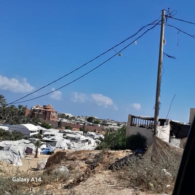 The image depicts an outdoor scene, likely a settlement or camp, situated in the city of Deir el-Balah, Palestinian Territories. The foreground is dominated by dry, rough terrain with sparse, dry vegetation. A prominent power pole stands off-center to the right, supporting multiple electrical wires that stretch across the frame against a clear blue sky with a few scattered clouds.

In the mid-ground, a densely populated area of temporary shelters, appearing to be white tents or fabric structures, stretches across the landscape. A lone palm tree stands among these shelters, and a blue car is visible. Further in the distance, a multi-story building with a reddish-brown roof and weathered walls is visible, surrounded by other structures and vegetation, suggesting a more established part of the settlement or a nearby town. The background shows a hilly landscape with scattered buildings and more vegetation, leading to a distant horizon.

The overall impression is one of a makeshift living arrangement, possibly a refugee or displaced persons' camp, in a somewhat arid environment. The presence of the power pole and wires indicates some level of infrastructure, while the abundance of tents suggests a temporary or emergency housing situation.