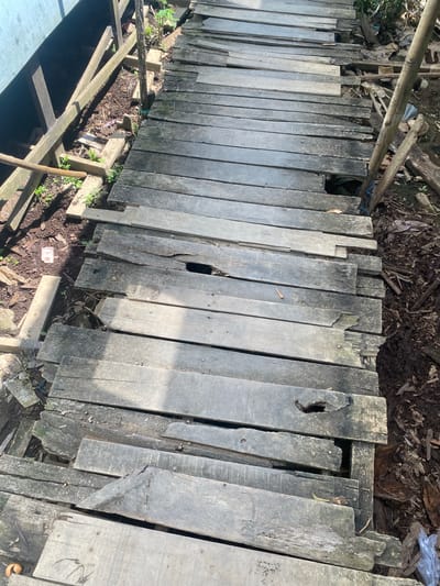 The image depicts a weathered wooden walkway in Berangas Timur, Indonesia. The walkway, constructed from aged, gray planks, shows signs of significant decay and damage. Several planks are broken, missing, or warped, creating gaps and uneven surfaces. The walkway is bordered by a dirt path with scattered debris, including leaves, plastic, and plant growth. Supporting wooden beams are visible beneath the walkway, some of which also appear damaged. The overall scene suggests an unmaintained or deteriorating pathway in an outdoor, possibly rural or semi-rural, environment.