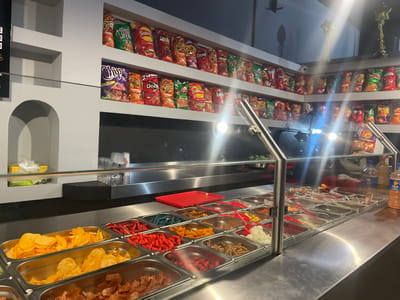 The image depicts the interior of a snack establishment, likely a chip bar, located in Fraccionamiento Los Heroes Queretaro, Mexico. The scene is dominated by a long, stainless steel counter with a clear glass sneeze guard extending across its length. Above the counter, recessed shelves integrated into a light grey wall display numerous bags of various branded chips, including Sabritas, Cheetos, Doritos, and Ruffles, arranged in multiple rows. An arched alcove on the left side of the wall contains a white napkin dispenser.
Below the glass guard, the stainless steel counter features a cold/hot bar configuration with multiple rectangular metal trays containing an assortment of snacks and toppings. Visible contents include large quantities of yellow potato chips (plain and ruffled), red chili-flavored snacks, blue-colored crunchy snacks, orange-colored chips, and various other savory toppings. Red plastic scoops are present in some of these trays. On the right side of the counter, a clear plastic bottle and an amber-colored condiment bottle are visible. Overhead lighting fixtures are reflected prominently in the glass and stainless steel surfaces, creating lens flares. No individuals are present in the image.