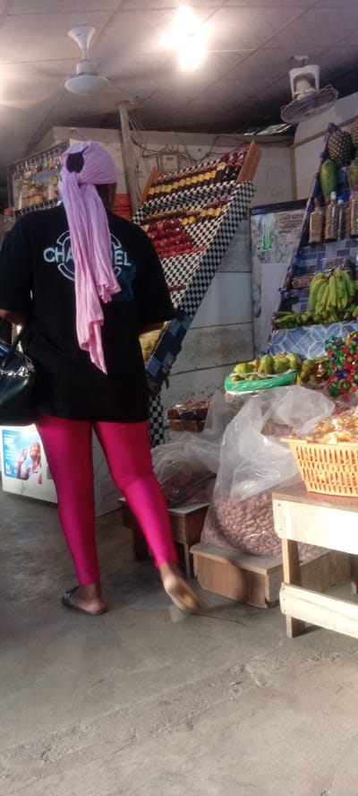 The image depicts an individual standing in what appears to be a market or store setting. The person is wearing a black t-shirt with "Chanel" printed on the back, bright pink leggings, and sandals. A pink headscarf is also visible. The person is standing near displays of fruits, vegetables, and other goods for sale. There are bags of peanuts on display. Overhead are ceiling fans and lights. The location is identified as Rijiyar Zaki, Nigeria.