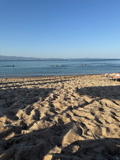 The image depicts a tranquil beach scene in Nea Potidea, Greece. The sun is low in the sky, casting long shadows across the sand, suggesting either early morning or late afternoon. The weather appears to be clear and warm, with a bright blue sky and calm, blue water.

The foreground is dominated by textured sand, marked with ripples and shadows, indicating it's a sandy beach. A few small pebbles are visible on the sand. In the mid-ground, the calm sea stretches out, with a few people enjoying a swim or wading in the water. One person is seen relaxing on a beach chair in the upper right portion of the image. A white sailboat is visible in the distance on the water, adding to the serene maritime atmosphere.

The background shows a hazy outline of land, likely a coastline with hills, characteristic of the Mediterranean landscape. The overall emotion conveyed is one of peaceful relaxation and enjoyment of a summer day at the beach. There is no visible text in the image.