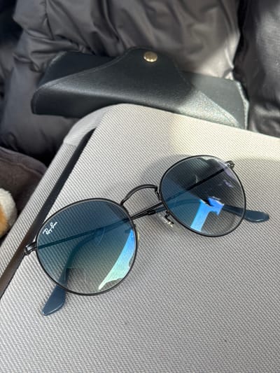 The image shows a pair of Ray-Ban sunglasses with black frames and blue-tinted lenses lying on a gray woven surface. Beside the sunglasses is a black leather case. Part of a black puffer jacket is visible in the background to the left. The location is likely an indoor setting, possibly in Langenfeld (Rheinland), Germany.