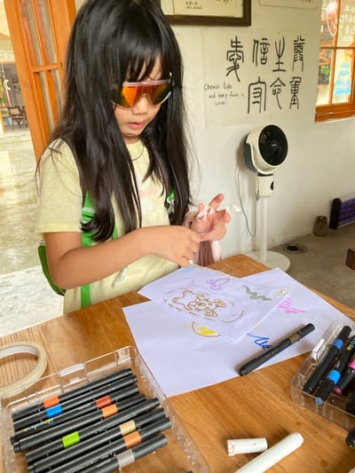 A young girl with long, dark hair and distinctive orange-tinted sunglasses is focused on drawing at a wooden table. She is wearing a pale yellow t-shirt with a green backpack strap visible. The table is scattered with art supplies, including a box of black markers with colored bands and white paper with drawings. To the left, a roll of tape is also visible. In the background, a white oscillating fan is mounted on a stand against a wall adorned with Chinese calligraphy and English text reading "Cherish life and keep faith in Love." The environment appears to be indoors, possibly in a classroom or workshop setting, with natural light filtering in from a doorway and a window. The overall scene suggests a moment of creative activity.