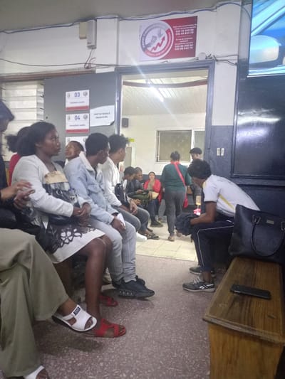 The image depicts a waiting area in a building located at coordinates -18.917 latitude and 47.517 longitude in the city of Antananarivo, Madagascar. There are several individuals, both men and women, seated on benches. Two women are wearing white clothing, one with a black patterned skirt, while a man in a white shirt and a man in a blue shirt sit with their legs extended in front of them. Two women sit further back, one in a red shirt and the other in a dark blue shirt. A male individual, in a white shirt and dark trousers, is seated on a bench to the right of the image, holding a black handbag. There is a wooden bench in the foreground, and a cell phone is lying on it. The walls are of a light grey colour, and the floor is tiled in a speckled grey colour. The building appears to be a government building, as there is a sign that reads "ZERO CORRUPTION," and there are also numbers on the wall. The entrance to the building is visible in the background. The lighting is dim.