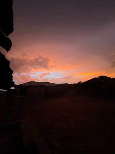 The image shows a dramatic, dusky sky at dusk or dawn, possibly in Iseyin, Nigeria, though details specific to Iseyin are not evident. The sky is characterized by a mix of purplish and orange hues, with cloud formations illuminated by the setting or rising sun. High-tension power lines cross the scene. The lower portion of the image is dominated by dark, silhouetted structures, likely buildings or parts of buildings. The lighting suggests that the buildings are casting long shadows, which obscures detail in these areas. The photograph appears to be taken from a low vantage point looking up at the sky.