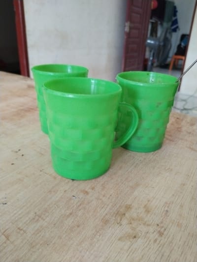 Three green plastic cups are arranged on a light-colored wooden surface. The cup in the foreground on the right is the most prominent, followed by a cup to its left, and a third cup partially visible behind the one on the left. The cups have a textured pattern of horizontal bands. A thin, dark object, possibly a utensil or wire, extends from the right cup towards the upper right corner of the image. The background is out of focus, showing a wall and what appears to be an open doorway with some objects visible within the room beyond. The scene is indoors, likely in a home or simple establishment. There is no indication of Meunasahtunong, Indonesia in the visual elements of the image.