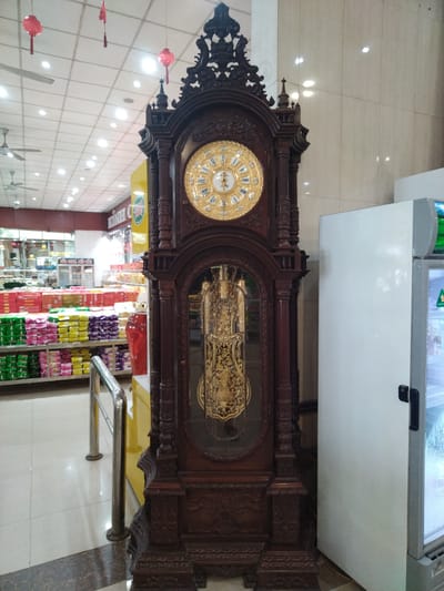 A floor-standing grandfather clock stands in a well-lit store with a tiled floor and drop ceiling. The clock is made of dark wood with ornate carvings and a gold-colored clock face. Behind the clock, shelves are stocked with colorful packaged goods, suggesting a grocery or retail store. To the right of the clock is a white refrigerator unit. The scene is indoors and appears to be within the confines of a retail establishment in Chu Văn An Ward, Vietnam.