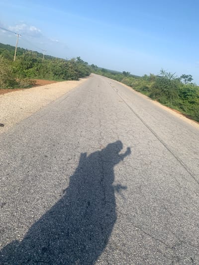 A long, empty asphalt road stretches into the distance, flanked by dry, grassy shoulders and sparse vegetation under a clear blue sky. The shadow of a person on a motorcycle is cast across the road, the rider's hand raised in a gesture that could be interpreted as waving. The scene appears to be in a rural or semi-rural area, possibly on the outskirts of Kardam C, Nigeria. The road is in fair condition, with some visible cracks. Utility poles with wires are present along the left side of the road, suggesting it is a maintained route. The overall impression is one of a quiet, sunny day in a landscape shaped by both natural elements and human infrastructure.