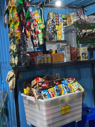 This media file depicts a retail setting, likely a small shop or market stall in Denasri Kulon, Indonesia, overflowing with various packaged snacks and food items. The scene is densely packed with colorful wrappers, creating a vibrant and somewhat chaotic display. On the left side, a large quantity of bagged snacks are neatly stacked or hung, featuring brands like "Energen," "Marin," and "Kopi." Towards the center, shelves are filled with more packaged goods, including boxes of cookies ("Oatstik," "Fullo," "Richoco") and milk cream snacks ("Kelapa Cream Susu Vanila"). A white plastic container on the right is filled with more snacks, including packets of "Rebo" crackers and "Nabati Wafer." The setting appears to be indoors, with a blue corrugated metal roof overhead and industrial-looking shelves. The lighting suggests it could be daytime or a well-lit interior. The overall impression is one of abundance and variety, typical of a local Indonesian convenience store or roadside stall. Visible text includes brand names, product descriptions like "Susu Vanila" (Vanilla Milk) and "Chocolate," and certifications like "HALAL." There's also a label on the white container indicating it's made from "100% MURNI PP COPOLYMER," "BPA FREE," and "FOOD CONTACT SAFE."